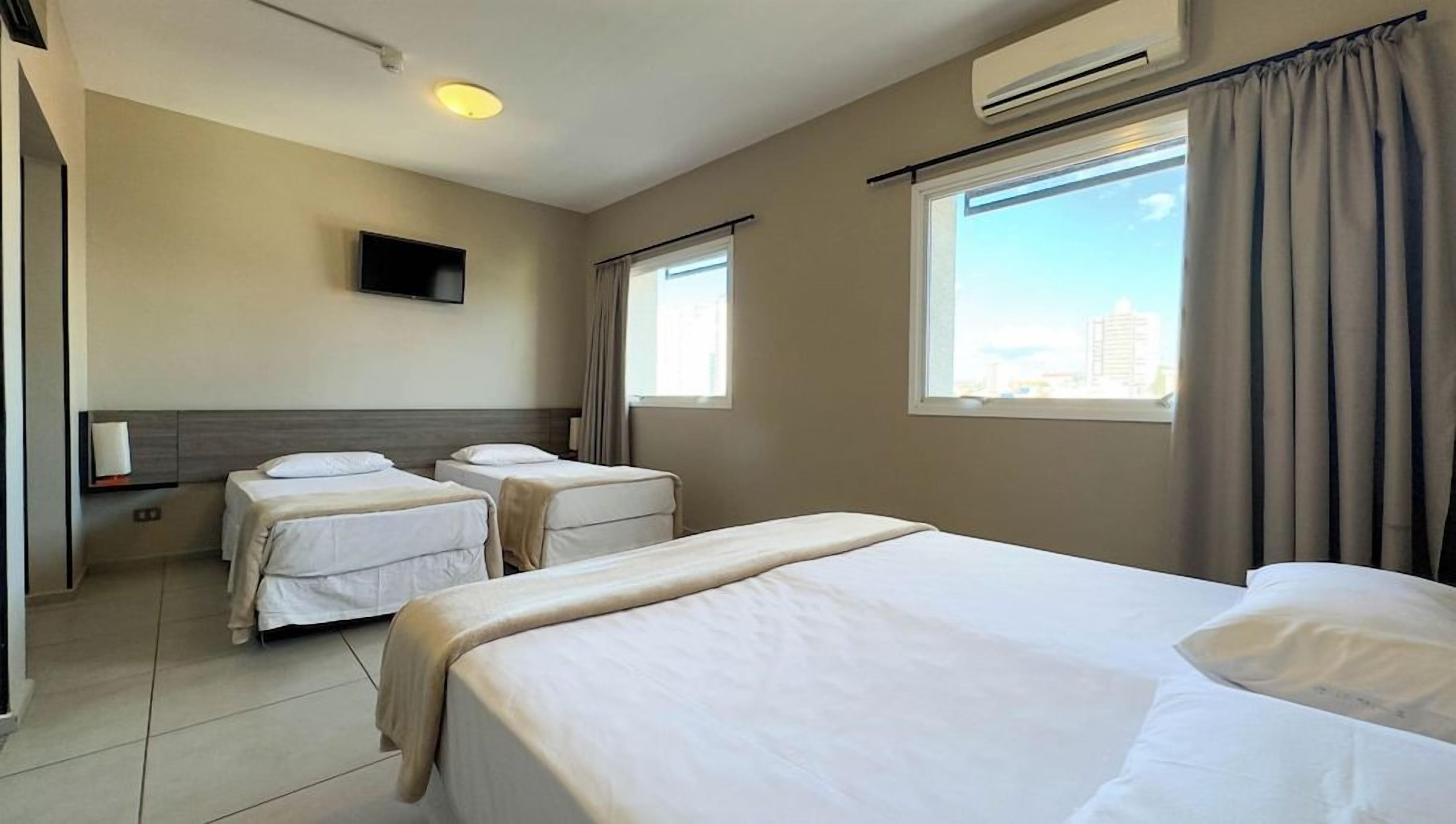 A hotel room with two single beds, a flat-screen TV mounted on the wall, and a large window with curtains.