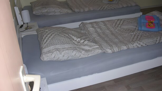 A room with two beds, each covered with a knitted blanket, a bedside table with a lamp, and a small toy on the right bed.