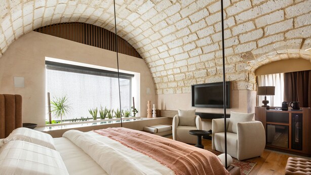 A bedroom with a large bed, a TV, a seating area with a sofa and armchairs, a window with plants, and a brick ceiling. - MBH | Tobacco Deluxe Room (Diso Spongano)