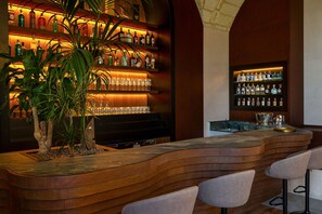 A bar area with a curved wooden counter, multiple shelves of liquor bottles, and a potted plant. - MBH | Tobacco Deluxe Room (Diso Spongano)