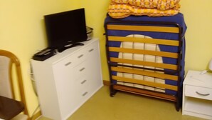 Una habitación con un aparador blanco, un televisor encima, una cuna azul y amarilla y una mesita de noche pequeña y blanca.