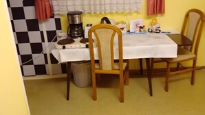 Un pequeño comedor con una mesa, sillas, cafetera y cubo de basura, además de una pared a cuadros.