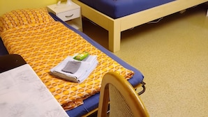 Una cama individual con colcha amarilla y naranja estampada, un cojín azul, una mesita de noche con lámpara y una silla con respaldo de malla.