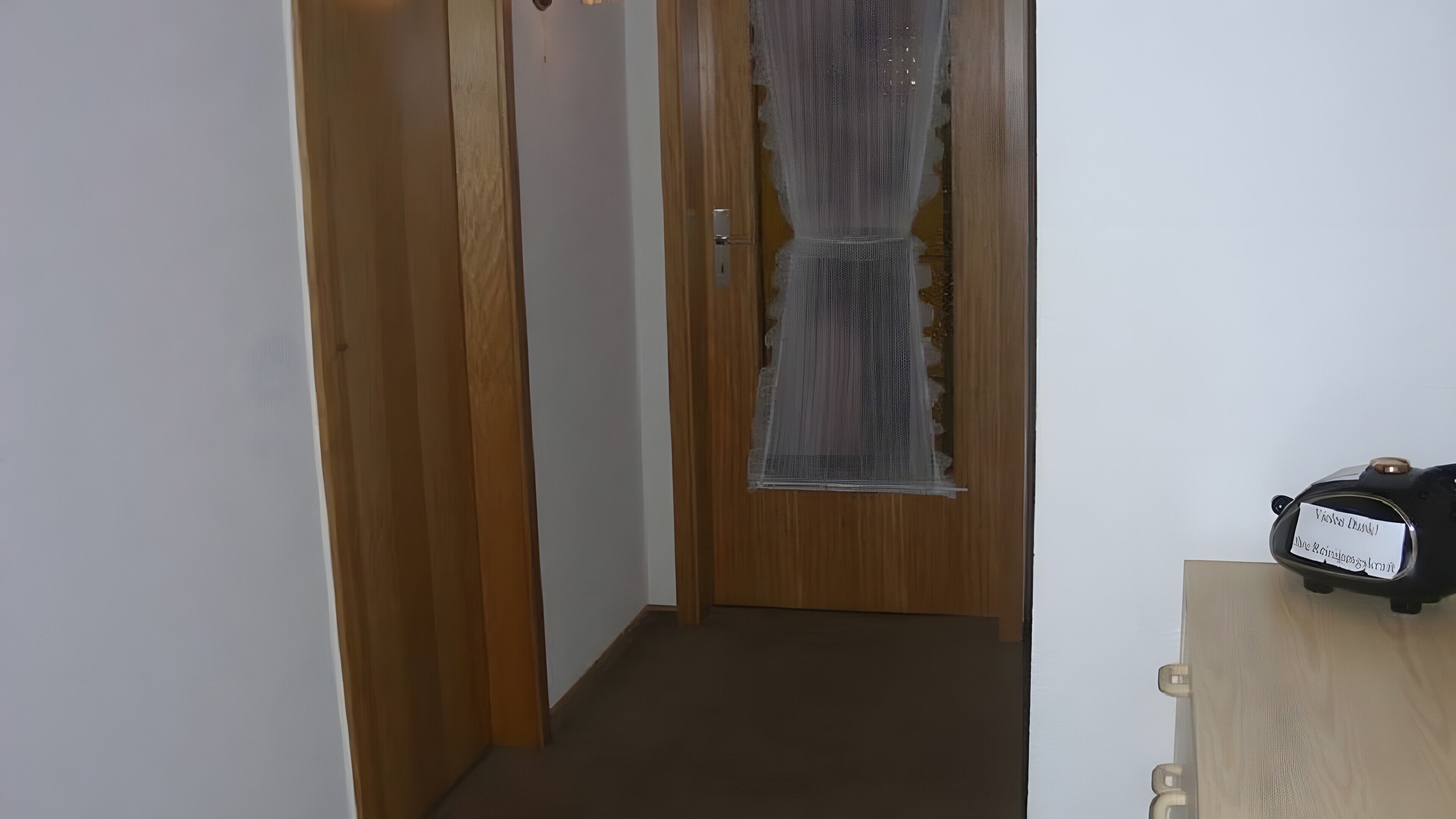 Um corredor com uma porta de madeira, uma janela com cortina e uma pequena mesa com um relógio.