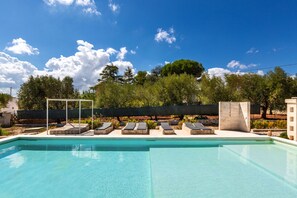 Une piscine avec des chaises longues, un espace de détente couvert et une vue sur les arbres et une clôture.
