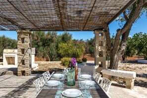An outdoor dining area with a pergola, stone tables, and chairs, surrounded by trees and a stone wall. - Villa Santa Lucia by Perle di Puglia (Ostuni)