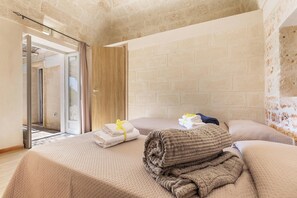 A hotel room with a brick wall, a bed with a beige bedspread, a wooden door, and a view through an open door. - Villa Santa Lucia by Perle di Puglia (Ostuni)