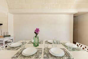 Une table à manger dressée avec des assiettes, des verres et des couverts, un vase rempli de fleurs et une petite pile de livres sur une table d’appoint.