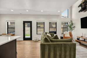 Ein modernes Wohnzimmer mit einer Couch, einem Couchtisch und einem Fernseher. Es gibt Fenster und eine Tür, die zu einem Außenbereich führt.
