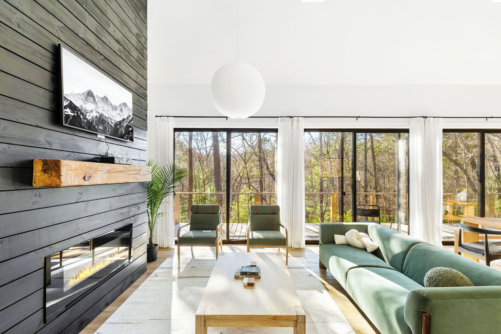 Un salon moderne avec une cheminée, un canapé vert et de grandes fenêtres donnant sur une forêt.