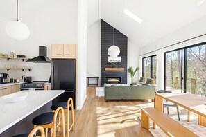A modern kitchen with a large island, a dining area with a wooden table and benches, and a living space with a sofa and fireplace. - BRAND NEW Modern Luxe Scandinavian Cabin with Hot tub/Fire Pit WIFI (Blue Ridge)