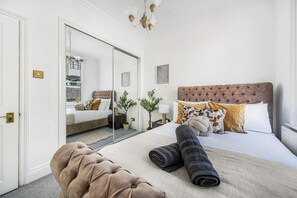 Ein Schlafzimmer mit einem großen Bett, einem Spiegel, einem gepolsterten Kopfteil und einem Nachttisch mit einer Lampe.