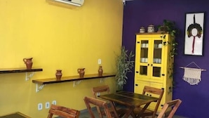 Ένας χώρος τραπεζαρίας με έναν κίτρινο τοίχο, έναν μωβ τοίχο που λειτουργεί ως σημείο έμφασης, μια κίτρινη βιβλιοθήκη με γυάλινες πόρτες και ένα τραπέζι με γυάλινη επιφάνεια, το οποίο πλαισιώνεται από ξύλινες καρέκλες.