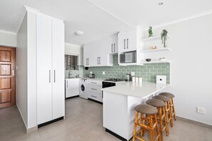 A modern kitchen with white cabinets, a microwave, a sink, and a bar stool area. - Rouxvilla (Jeffreys Bay)