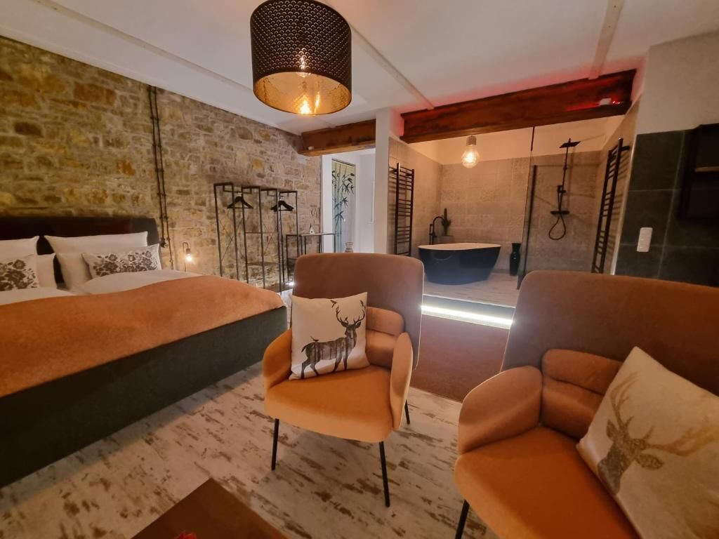 A bedroom with a large bed, two armchairs, a hanging light fixture, and a stone wall.