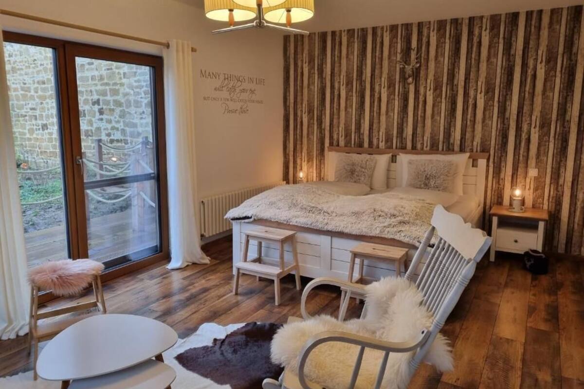 A bedroom with a large bed, a wooden headboard, a small bedside table, a chair, a window with curtains, and a wall with a quote.
