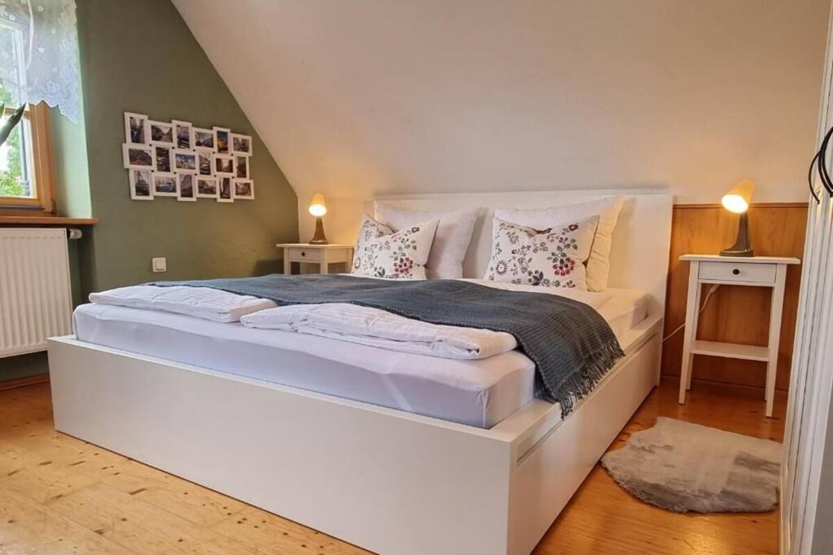 A bedroom with a white bed, a small bedside table, a lamp, and a wall with photos.