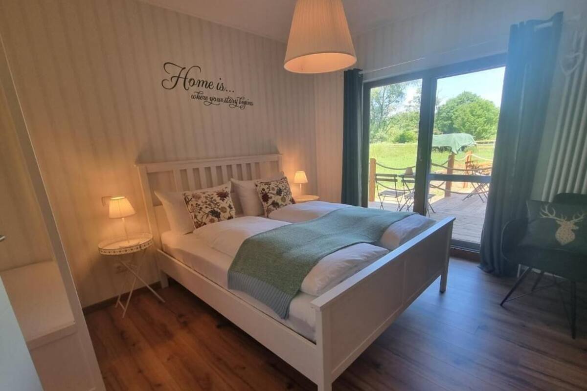 A bedroom with a white bed, a bedside table, a lamp, and a view of an outdoor area with a table and chairs.