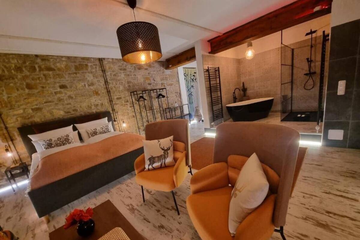 A modern room with a bed, two armchairs, a coffee table, a stone wall, and a bathroom with a bathtub and shower.