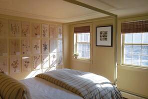 Une chambre avec un lit, des reproductions botaniques encadrées accrochées au mur, une fenêtre donnant sur un beau paysage et une petite plante en pot.