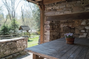 Une table en bois rustique, ornée d’un pot de fleurs, adossée à un mur de pierres. À l’extérieur, on aperçoit un mur de pierres et des poutres en bois, ainsi qu’un banc en bois.