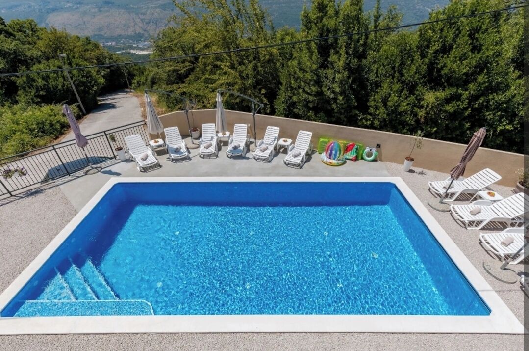 A rectangular pool with clear blue water, surrounded by white lounge chairs and umbrellas, set against a backdrop of trees and a mountainous landscape.