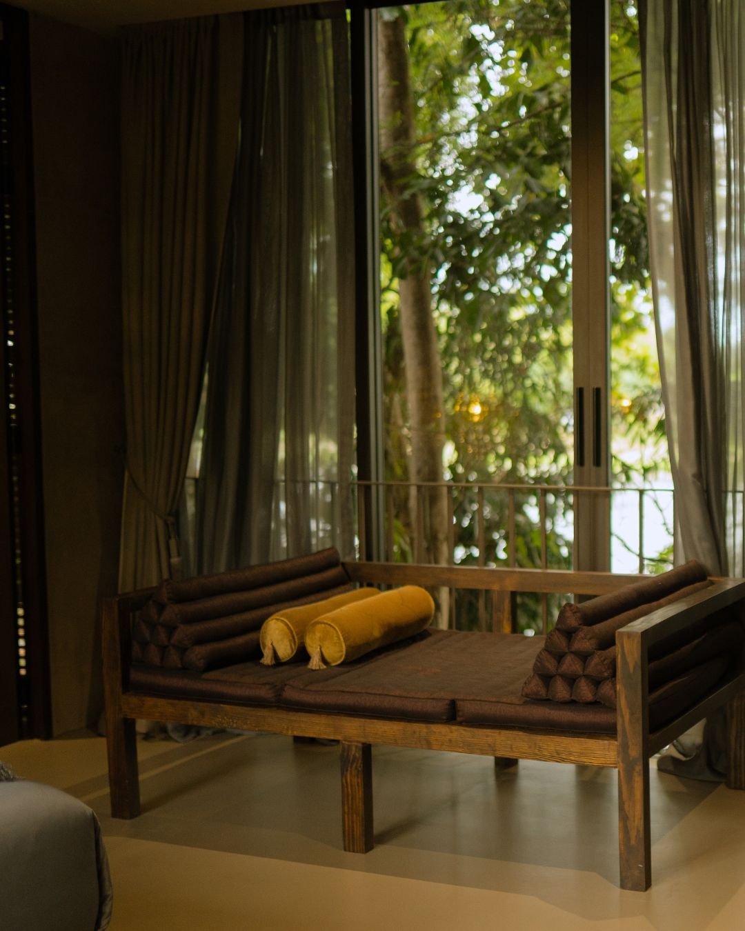 A wooden bed with rolled-up blankets and pillows, positioned in a room with large windows and curtains.