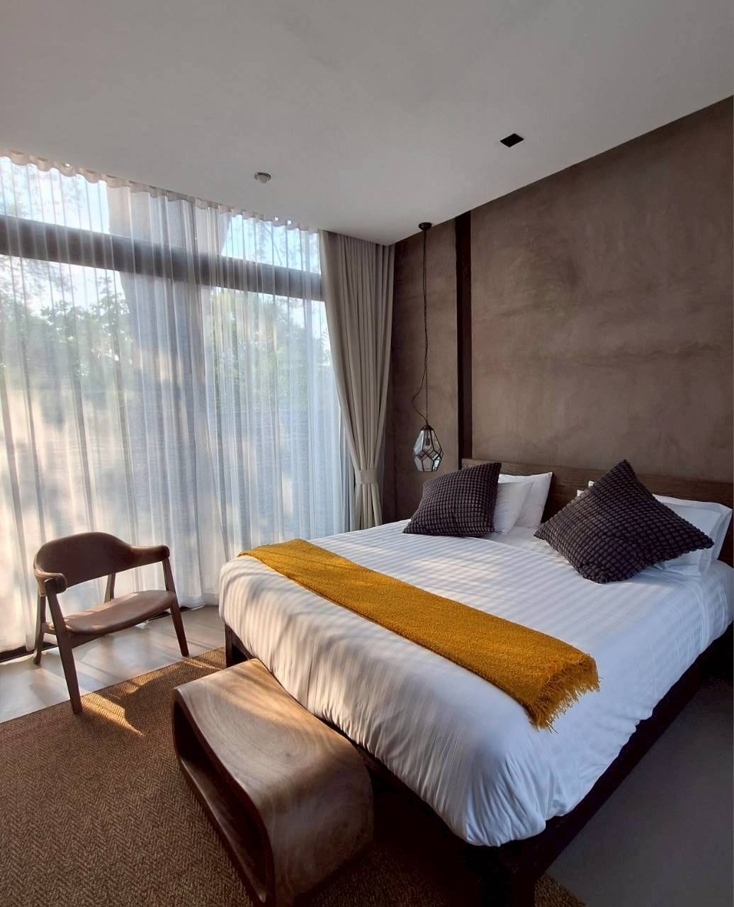 Une chambre moderne avec un grand lit, un banc en bois et une vue sur les arbres à travers des rideaux transparents.