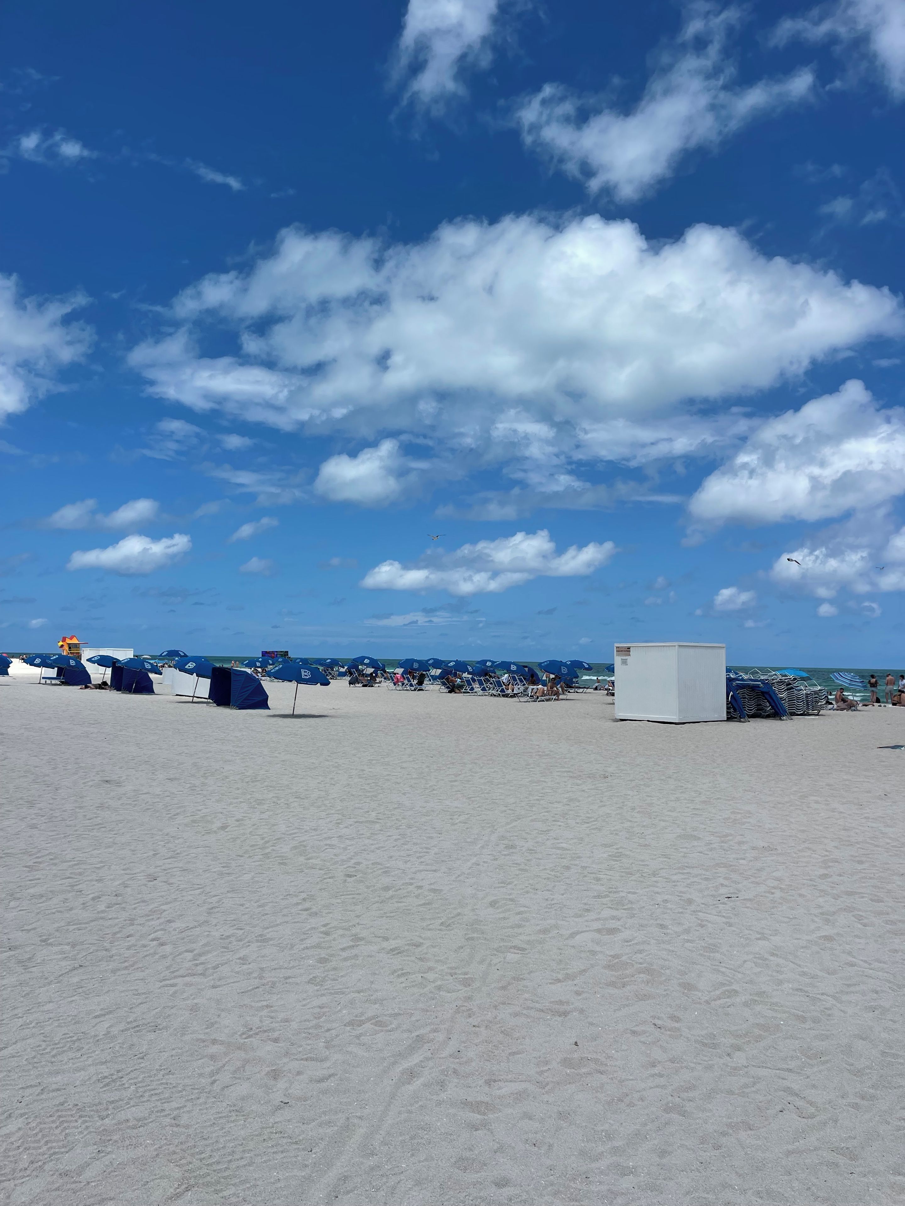 A beach with umbrellas and beach chairs, a lifeguard tower, and the ocean in the background.