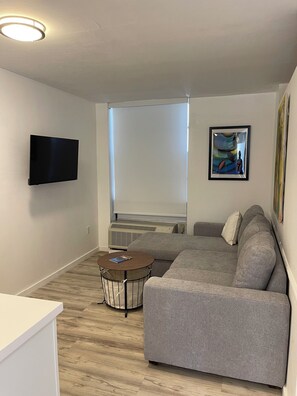 Ein modernes Wohnzimmer mit grauem Sofa, einem flachbildfernseher an der Wand, einem runden Couchtisch mit einem Buch und einem großen Fenster mit Jalousien.