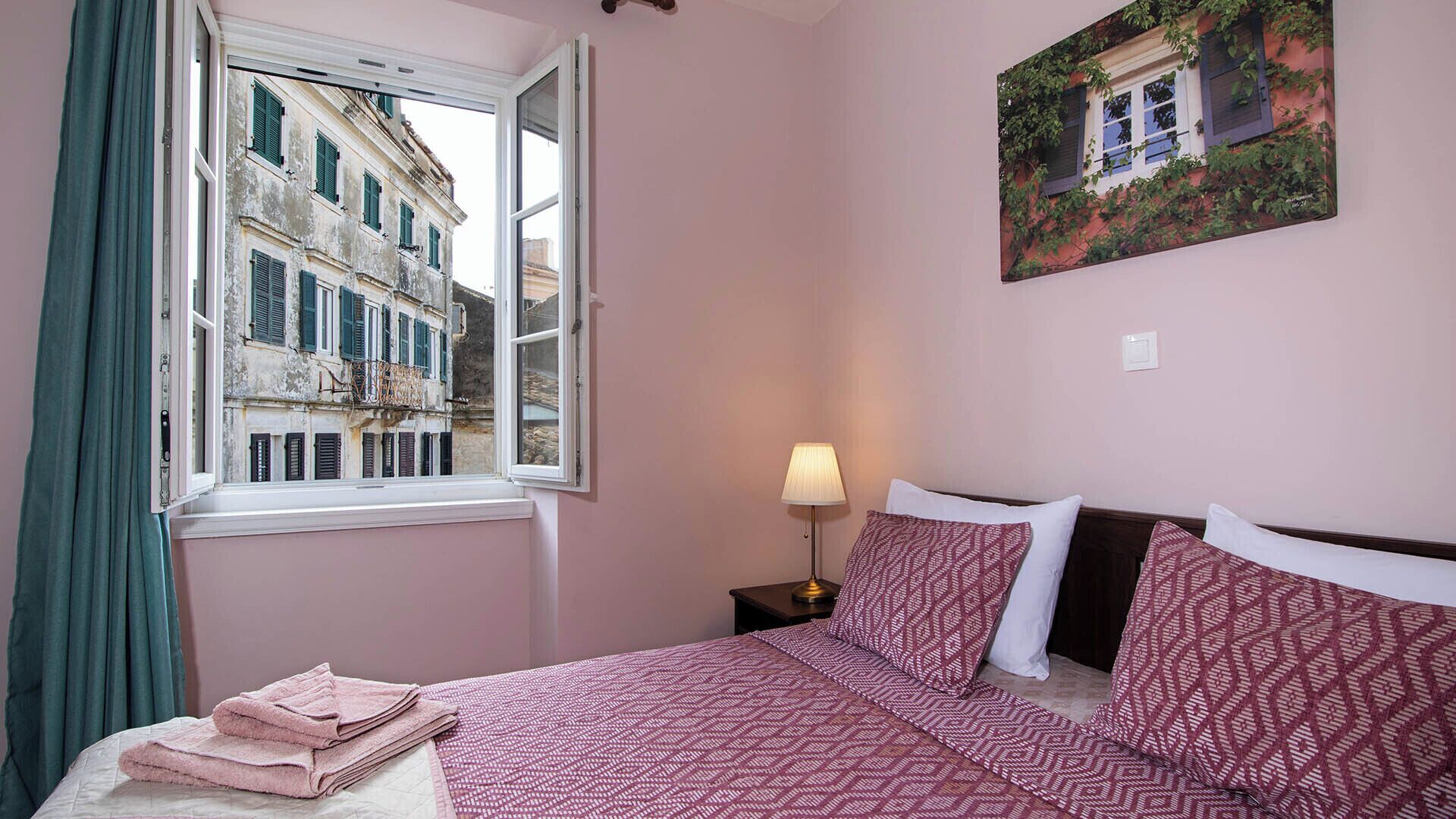 Ein Schlafzimmer mit Bett, Nachttisch, Lampe, Fenster mit Blick auf Gebäude und einem Bild an der Wand.
