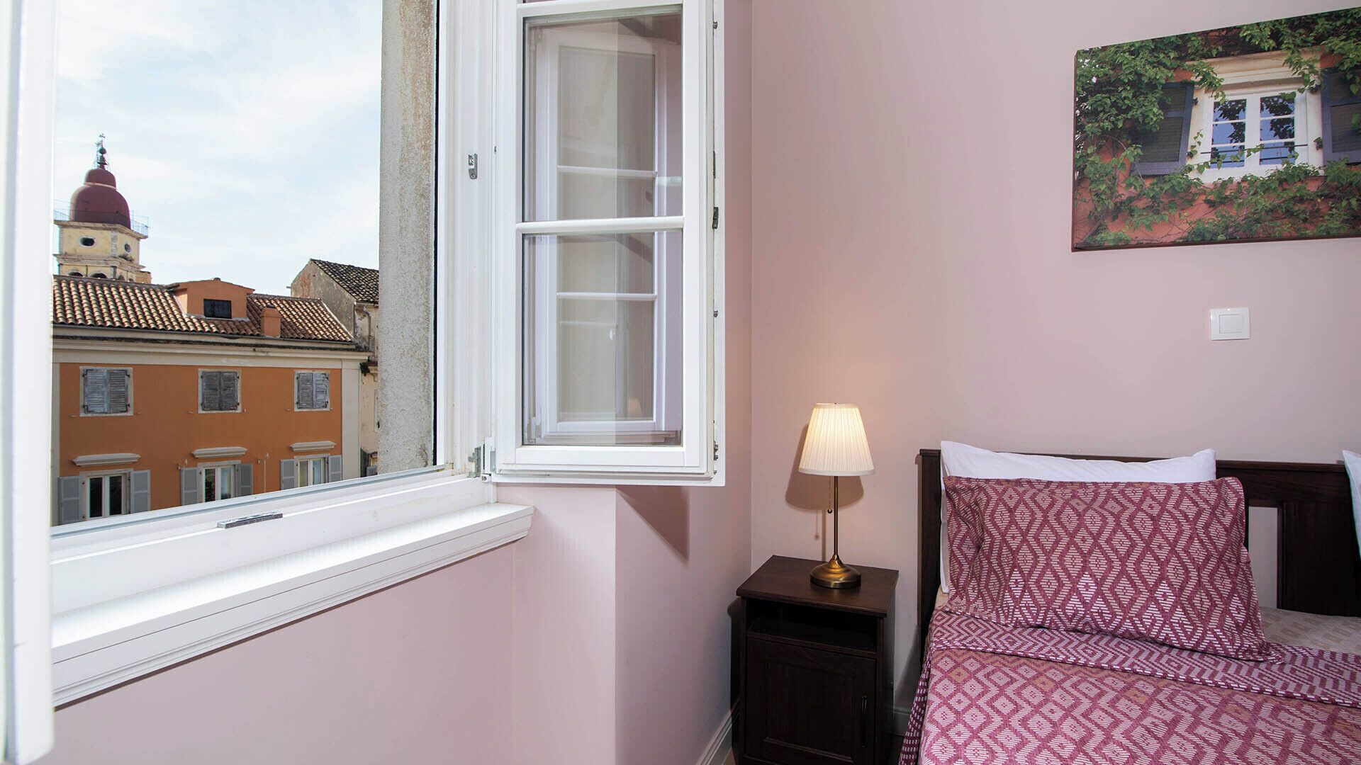 Une chambre avec une fenêtre, une lampe de chevet, un lit avec une literie à motifs et un tableau au mur.
