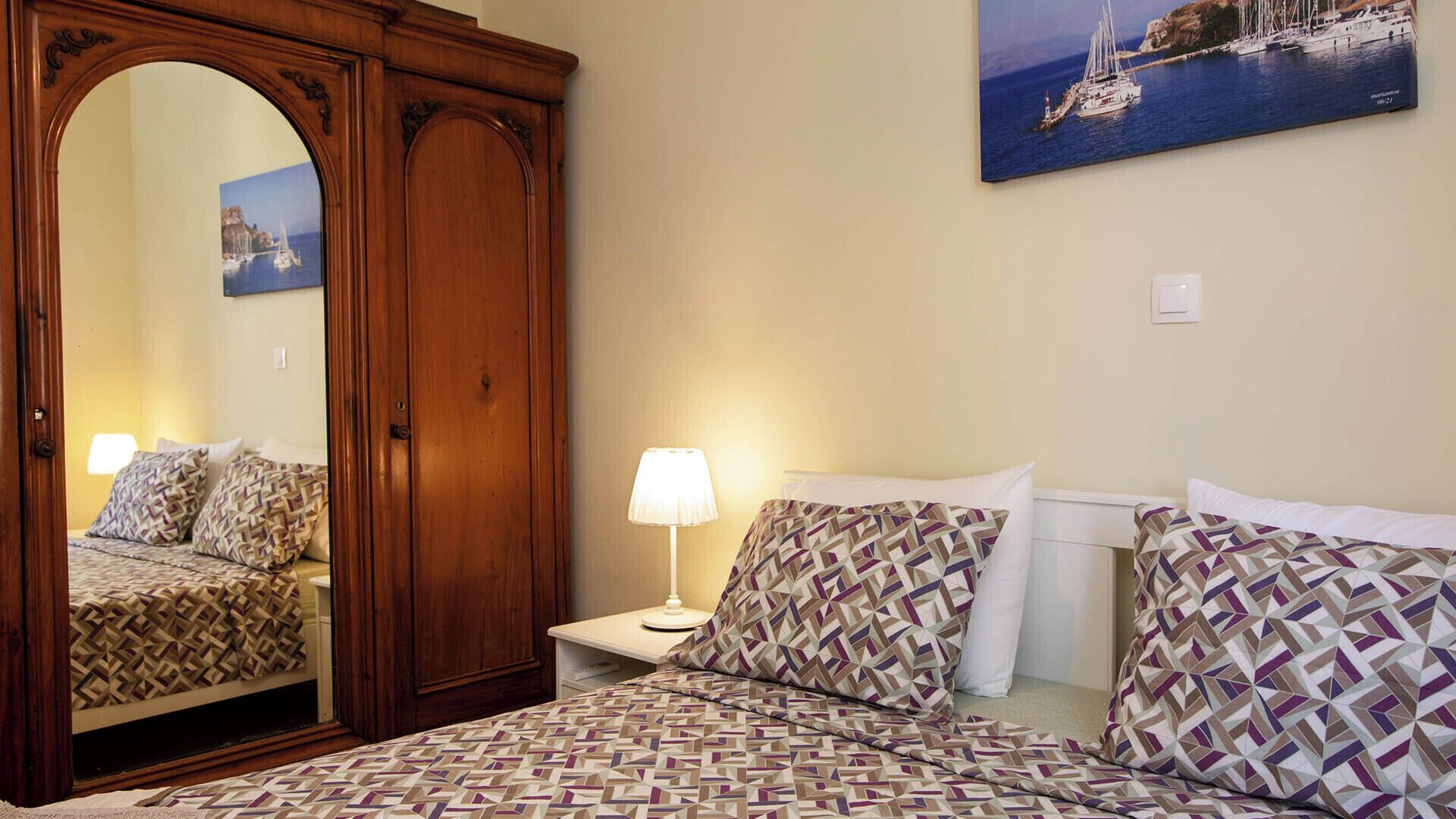 Quarto com cama, almofadas estampadas, mesa de cabeceira com abajur, guarda-roupa de madeira e quadro na parede.