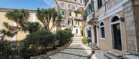 A stone-paved courtyard with buildings, some with balconies, and greenery.