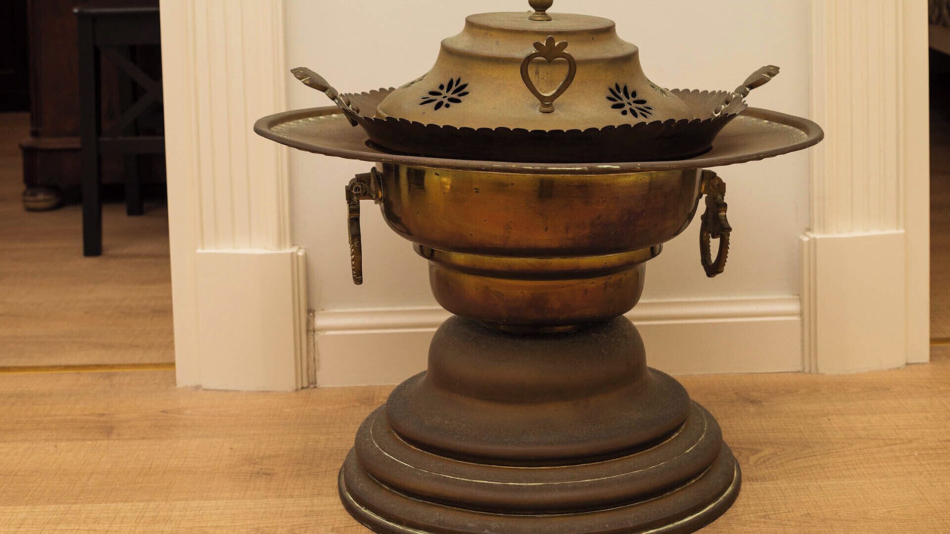 A brass stove with a decorative lid and handles, placed on a wooden floor.