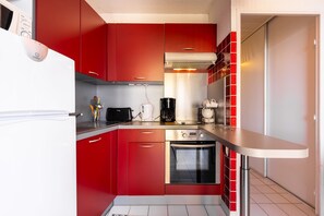 A compact kitchen with red cabinets, a white refrigerator, a coffee maker, and a built-in oven. - Valras-Plage  Appartement avec Vue Mer (Valras-Plage)