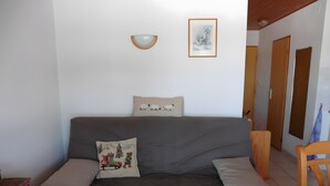 Habitació amb un sofà gris, dos coixins, una làmpada de paret, un quadre amb marc i una porta.