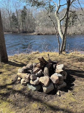 Luonnonkauniissa ympäristössä oleva nuotiopaikka, jonka ympärillä on kiviä, joki ja puita.