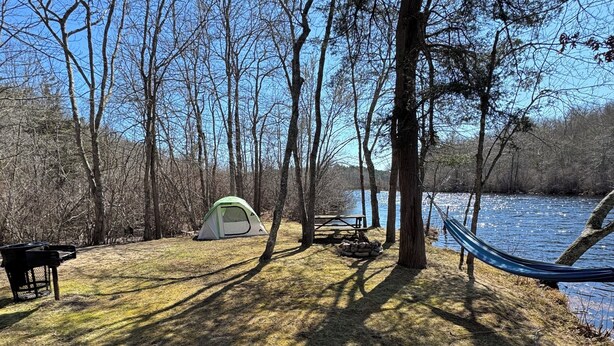 Un camping con tienda de campaña, mesa de picnic, hamaca y terraza de madera con vista a un cuerpo de agua.