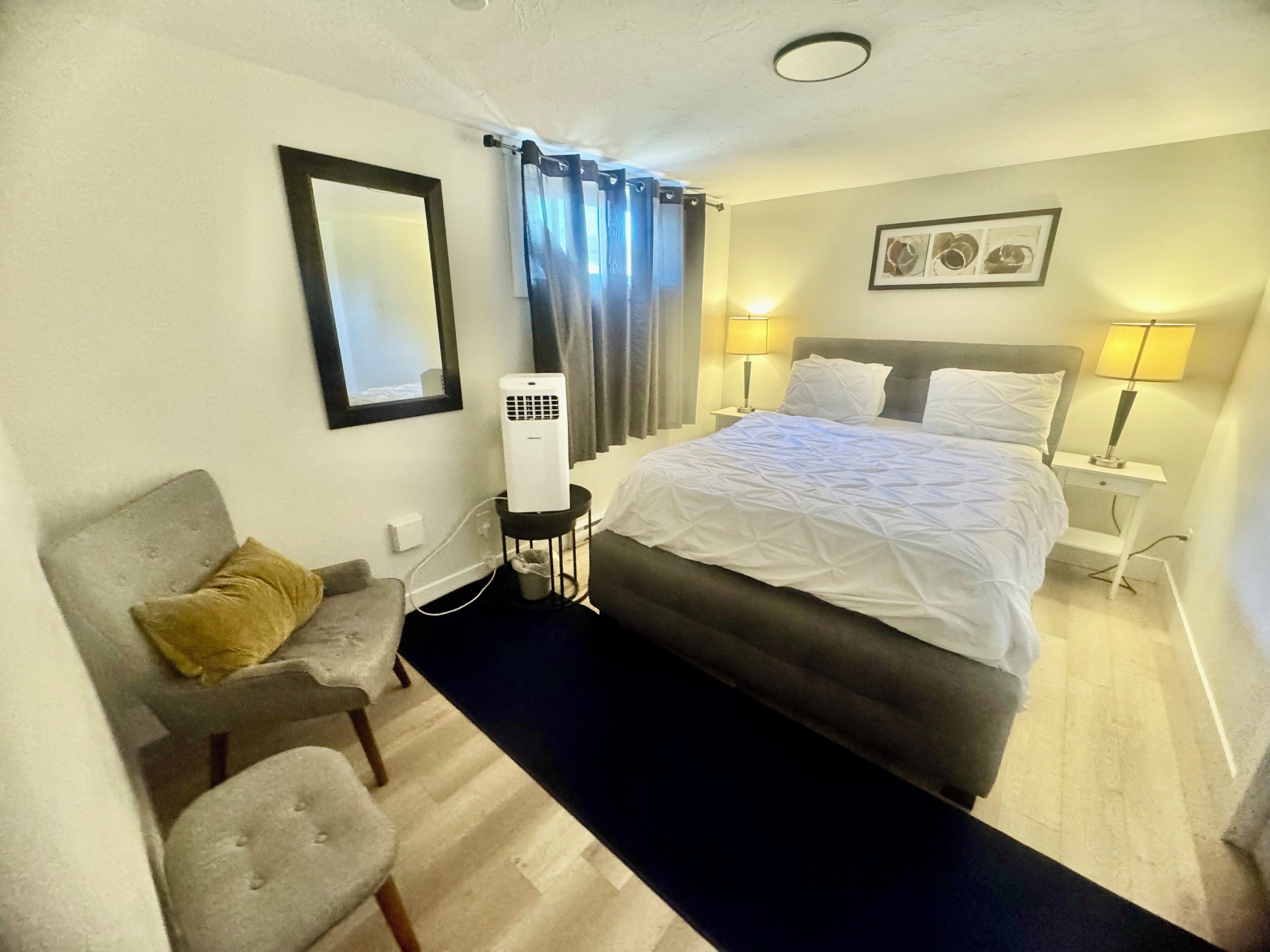 Una habitación de hotel con cama, lámparas de noche, un calentador, una silla, una mesita y un espejo.
