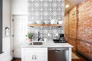 A kitchen with white cabinetry, a marble countertop, a brick wall, and a patterned backsplash. - Vibrant Tennyson Victorian | 96 Walk Score | 30Day (Denver)