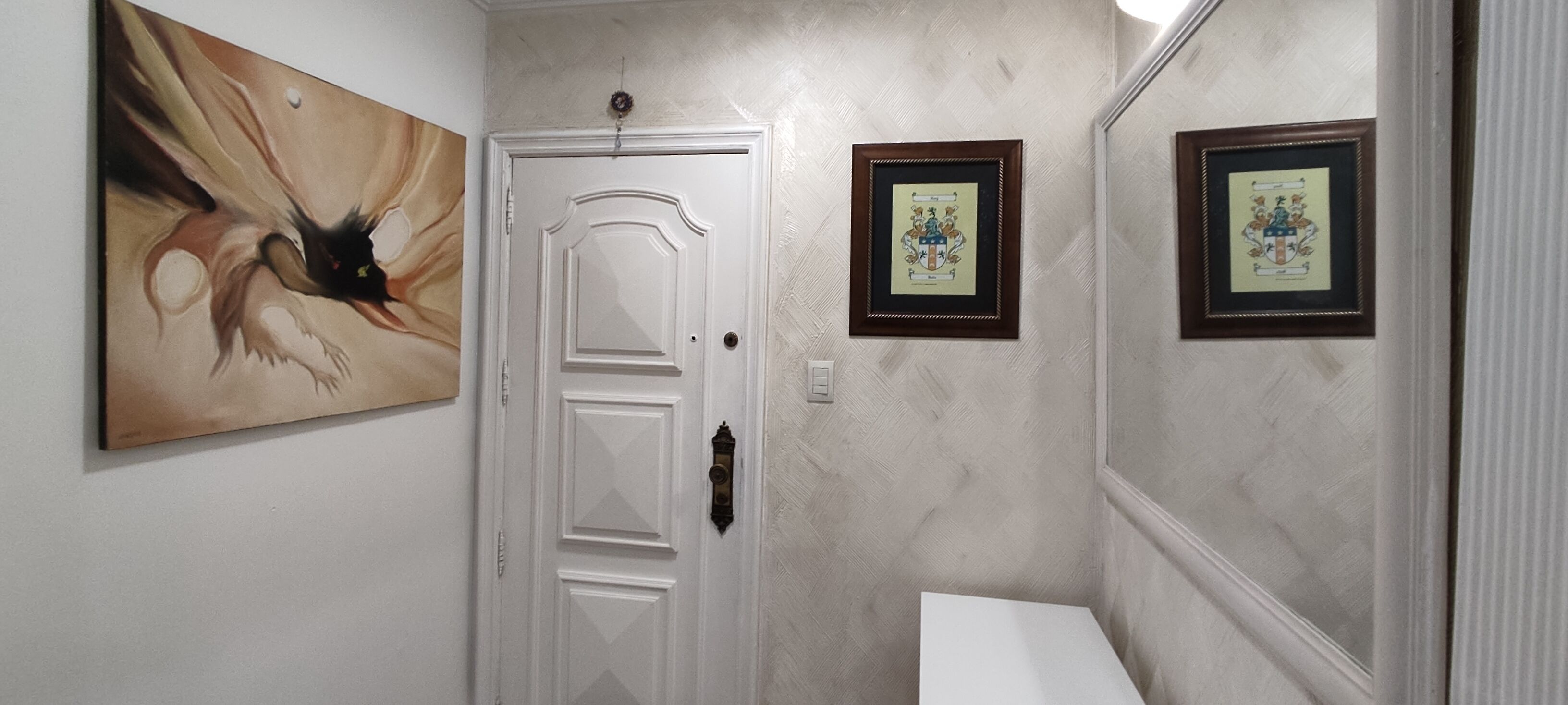 Un pasillo con una puerta blanca, un escudo enmarcado en la pared y un cuadro.