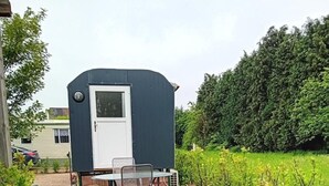 Een klein, donker gebouw buiten met een deur en ramen, een tafel met stoelen, en een groen gazon met een stoel.