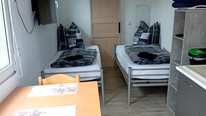 Een kleine kamer met twee bedden, een tafeltje, een stoel, een aan de muur bevestigde televisie en een houten deur.