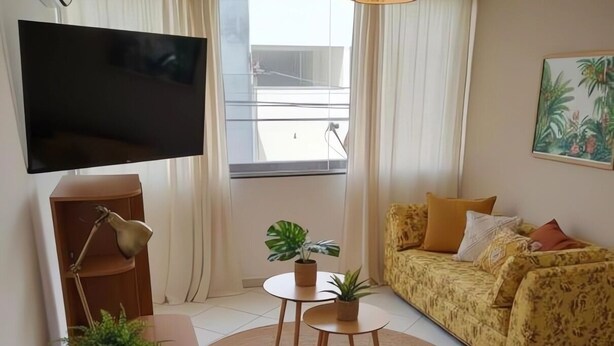 Hotellihuone, jossa on sänky, sohva, lamppuvarusteinen työpöytä, televisio, ikkuna verhoilla ja huonekasvi.