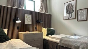 Hotellihuone, jossa on kaksi sänkyä, puupanelilla verhoiltu seinä, pieni työpöytä laatikoineen, kehystetty taulu ja seinään kiinnitetty lamppu.