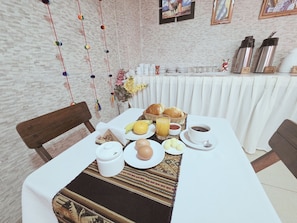 Una mesa de desayuno con mantel blanco, una cesta de mimbre, un plato de huevos, un vaso de jugo de naranja, una taza de café y un termo.