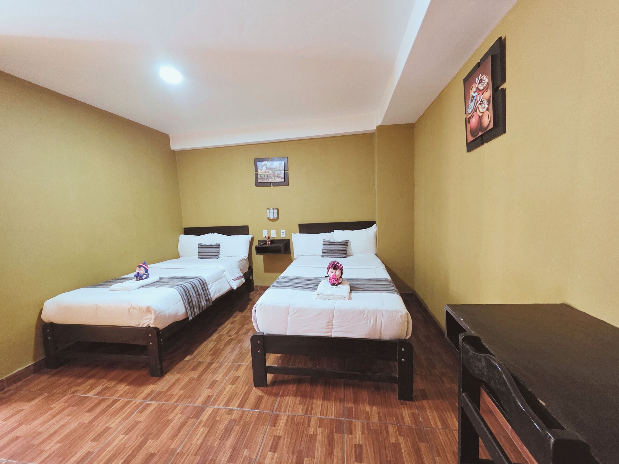 A hotel room with two beds, a small table, a wall-mounted picture, and a ceiling light.