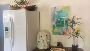 Una cocina con un refrigerador blanco, un banco de madera, una cesta tejida, una planta en maceta y un cuadro abstracto colorido en la pared.