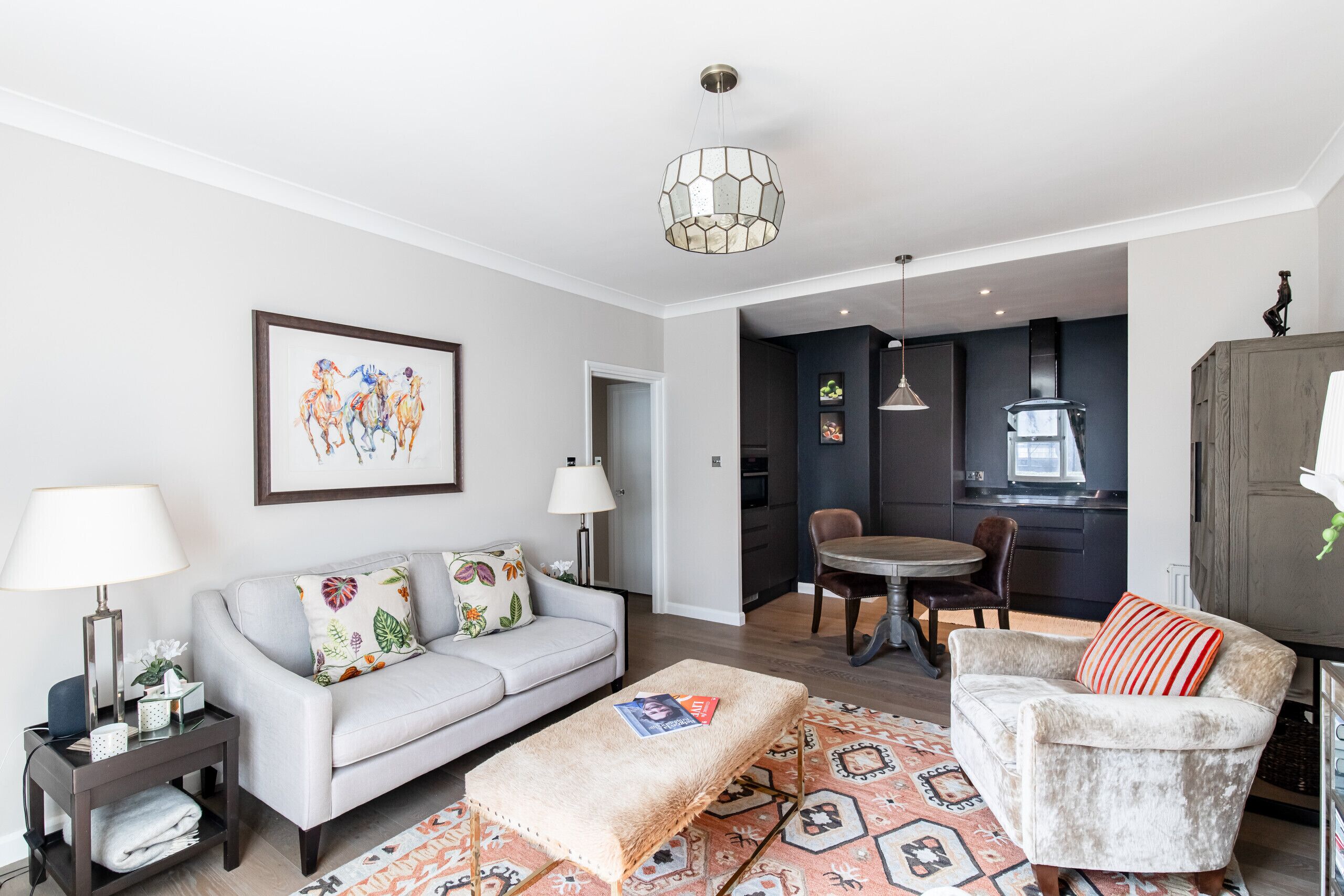 Un salon moderne avec un canapé, un fauteuil et un pouf, un coin repas avec une table et des chaises, et une cuisine en arrière-plan.